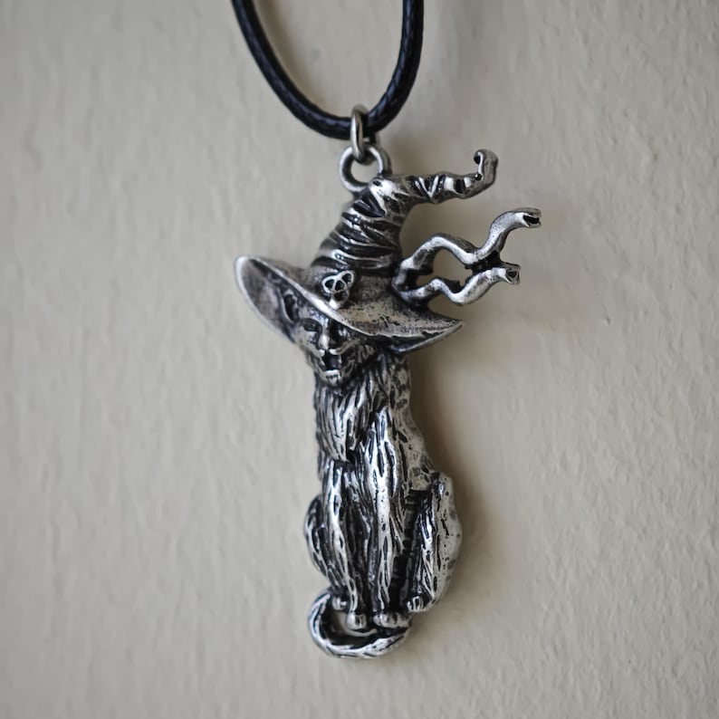 May include: A silver-colored pendant necklace featuring a cat wearing a witch's hat. The cat is seated, with a long, fluffy coat and a curled tail. The witch hat has a skull and a curly design. The necklace has a black cord.