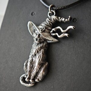 May include: A silver-colored pendant necklace featuring a cat wearing a witch's hat. The cat is seated, with detailed fur and facial features. The hat has a skull and wavy accents. The pendant hangs from a black cord.
