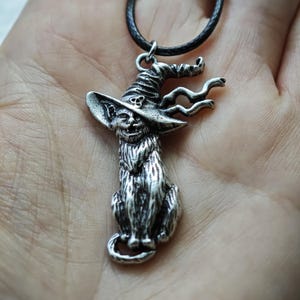 May include: A silver-colored pendant necklace featuring a cat wearing a witch's hat. The cat is seated with a long, detailed body and a smiling face. The witch hat has a skull on it and the necklace has a black cord.