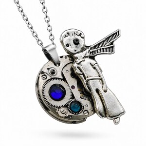 Peut inclure: Un collier pendentif argenté composé d'un mécanisme de montre circulaire avec deux cristaux bleus incrustés. Une figurine métallique représentant une personne portant une écharpe est fixée sur le côté de l'assemblage d'engrenages, sur une chaîne.