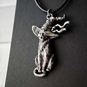 May include: Silver-toned pendant necklace featuring a cat wearing a witch's hat. The cat is seated, with detailed fur and a whimsical hat design. The necklace has a black cord.