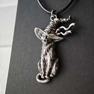 May include: Silver-toned pendant necklace featuring a cat wearing a witch's hat. The cat is seated, with detailed fur and a whimsical hat design. The necklace has a black cord.