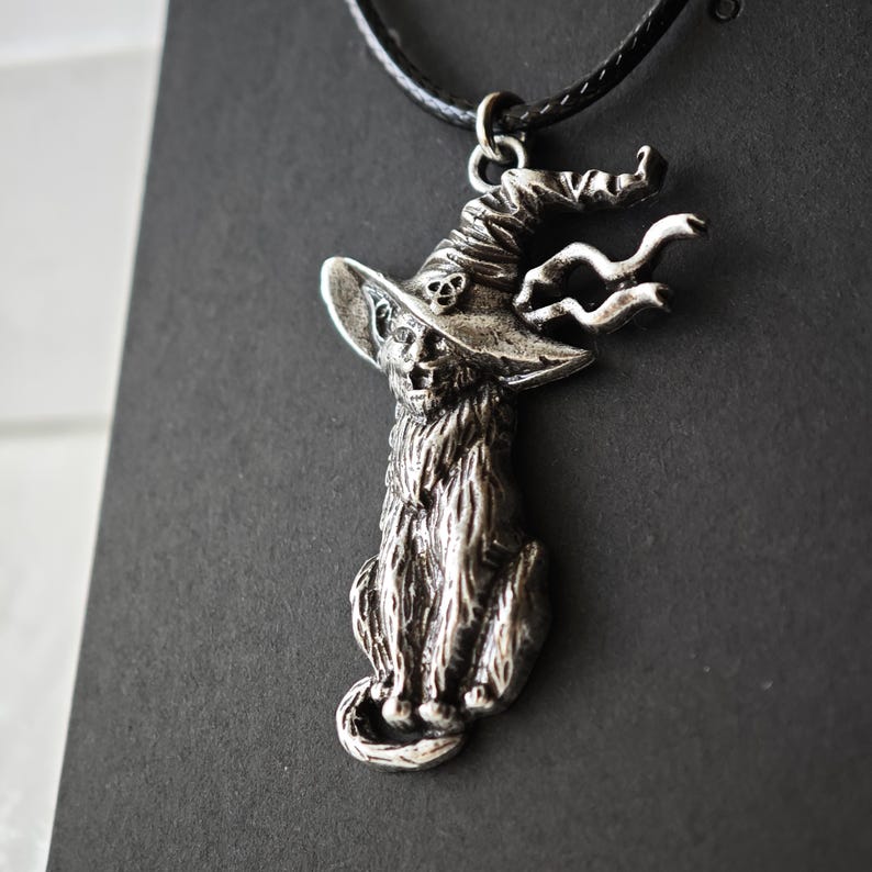 May include: Silver-toned pendant necklace featuring a cat wearing a witch's hat. The cat is seated, with detailed fur and a whimsical hat design. The necklace has a black cord.