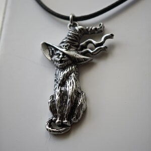 May include: A silver-toned pendant necklace featuring a cat wearing a witch's hat. The cat is seated with its tail curled. The hat has a skull detail. The pendant hangs from a black cord.