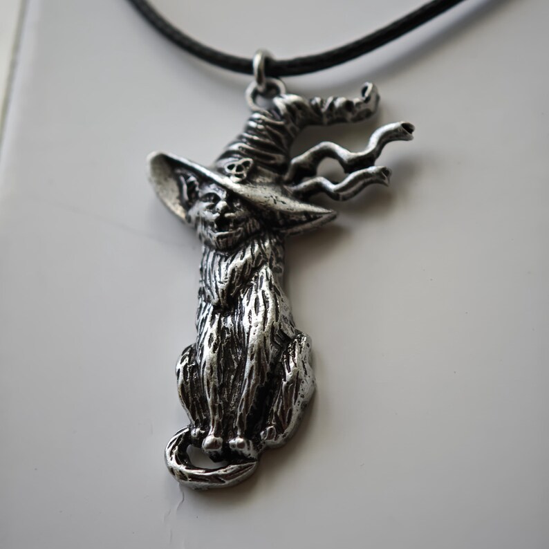 May include: A silver-toned pendant necklace featuring a cat wearing a witch's hat. The cat is seated with its tail curled. The hat has a skull detail. The pendant hangs from a black cord.