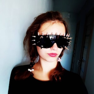 Op de afbeelding: Zwarte zonnebril met zilverkleurige spikes langs de boven- en zijkanten. De zonnebril heeft een rechthoekige vorm. De persoon die hem draagt heeft bruin haar in vlechten en draagt een zwarte top.