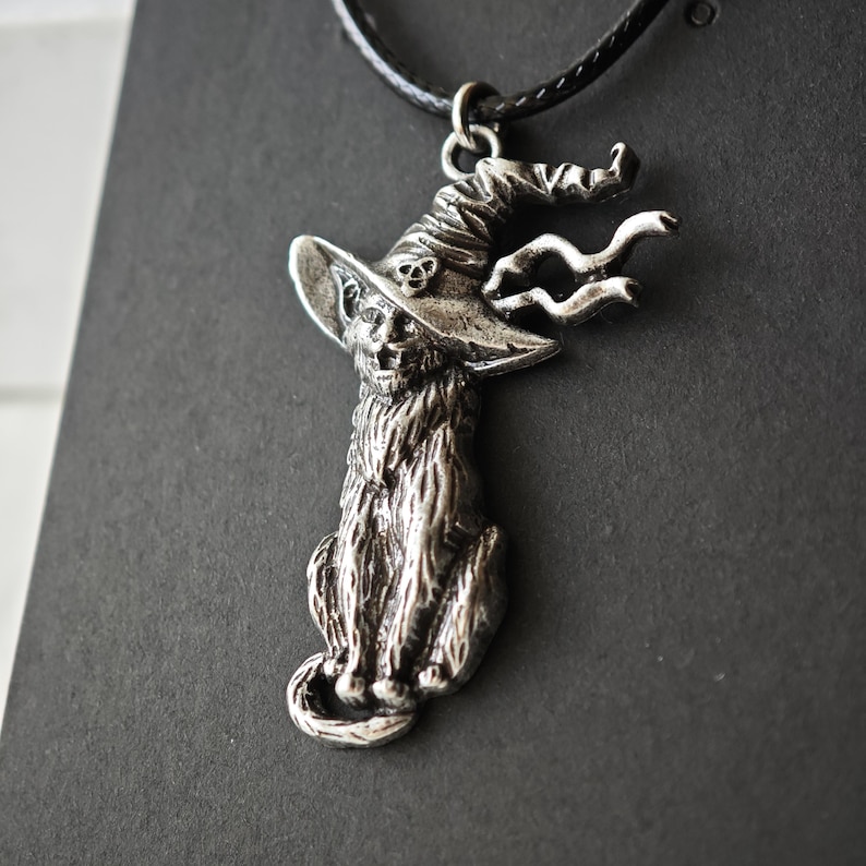 May include: A silver-toned pendant necklace featuring a cat wearing a witch's hat with a skull detail. The cat is seated, with detailed fur texture. The pendant hangs from a black cord, perfect for Halloween or gothic-themed jewelry.