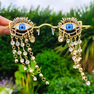 May include: Gold-coloured eye-shaped sunglasses with blue irises and crystal accents. The frames feature dangling crystal chains and a decorative sunburst design around the eye. The sunglasses are held against a blurred green background.