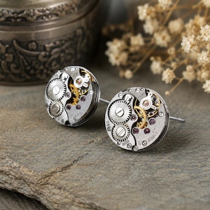 May include: A pair of circular stud earrings crafted from repurposed silver-toned watch movements. Each earring features exposed metallic gears, small brass-coloured cogs, and tiny purple jewel bearings arranged within a round mechanical frame.