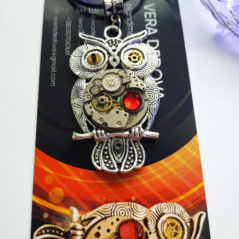 May include: Silver-toned owl pendant necklace with intricate clockwork details and a red gemstone. The owl's eyes are gold-colored. The pendant hangs on a black cord. The background includes a business card with contact information.