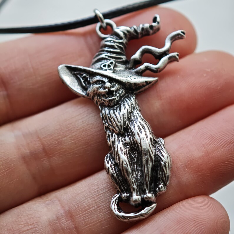 May include: A silver-toned pendant necklace featuring a cat wearing a witch's hat. The cat is seated, with a long, textured fur design. The witch hat has a curled brim and a Celtic knot. The necklace is on a black cord.