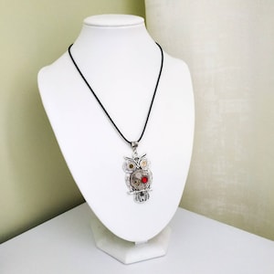 May include: A silver-toned owl pendant necklace with a black cord. The owl pendant features intricate mechanical gears, a red gem, and detailed feather designs. The necklace is displayed on a white bust.