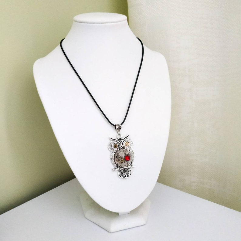 May include: A silver-toned owl pendant necklace with a black cord. The owl pendant features intricate mechanical gears, a red gem, and detailed feather designs. The necklace is displayed on a white bust.