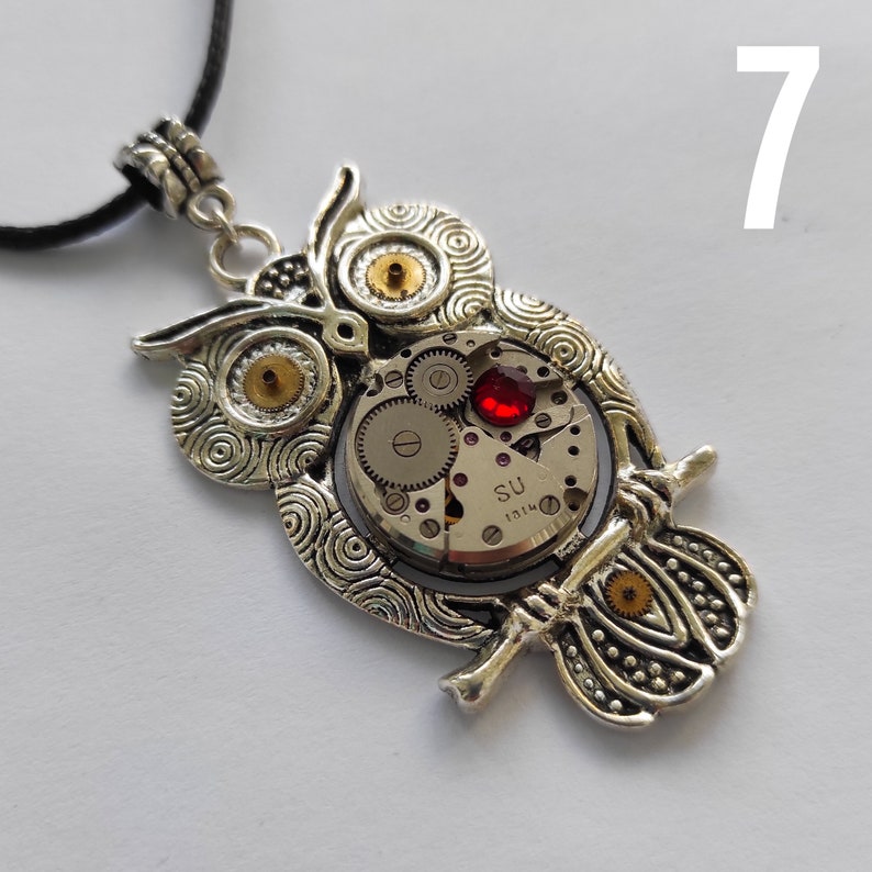 May include: Silver-toned owl pendant necklace with intricate details. The owl's body features a watch mechanism with visible gears and a red jewel. The pendant hangs from a black cord, creating a unique steampunk accessory.