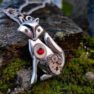 Puede incluir: Un collar con un colgante de zorro plateado con una joya roja y detalles intrincados de relojería. El colgante está sentado sobre una roca cubierta de musgo.