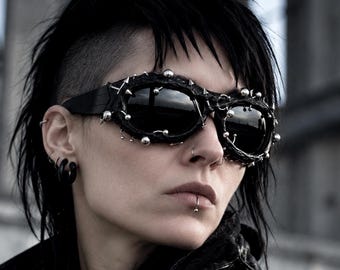 May include: Black sunglasses featuring a textured, rugged frame adorned with various silver-toned metal studs, spikes, and small rings. The lenses are dark and reflective, and the overall design incorporates multiple metallic embellishments.