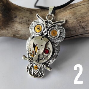 May include: Silver-toned owl pendant necklace with a steampunk design. The owl features intricate details, including watch gears for eyes and a red gem. The pendant hangs from a black cord.