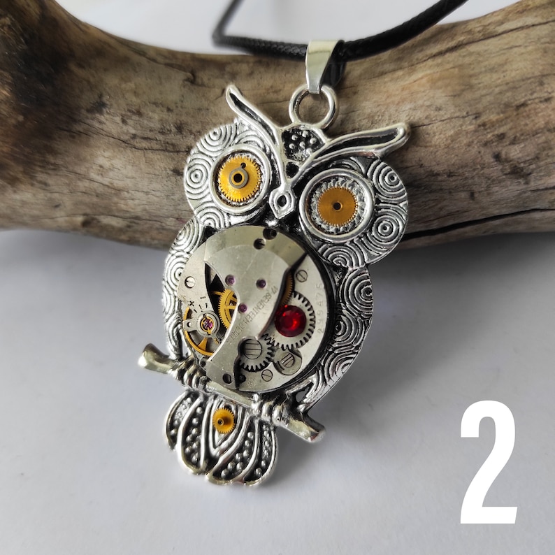 May include: Silver-toned owl pendant necklace with a steampunk design. The owl features intricate details, including watch gears for eyes and a red gem. The pendant hangs from a black cord.