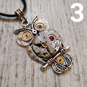 May include: Silver-toned owl pendant necklace with a black cord. The owl design incorporates watch gears and a red gem. The pendant is approximately 2 inches in height and 1.5 inches in width.