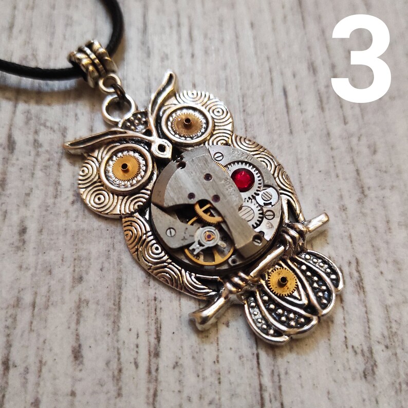 May include: Silver-toned owl pendant necklace with a black cord. The owl design incorporates watch gears and a red gem. The pendant is approximately 2 inches in height and 1.5 inches in width.