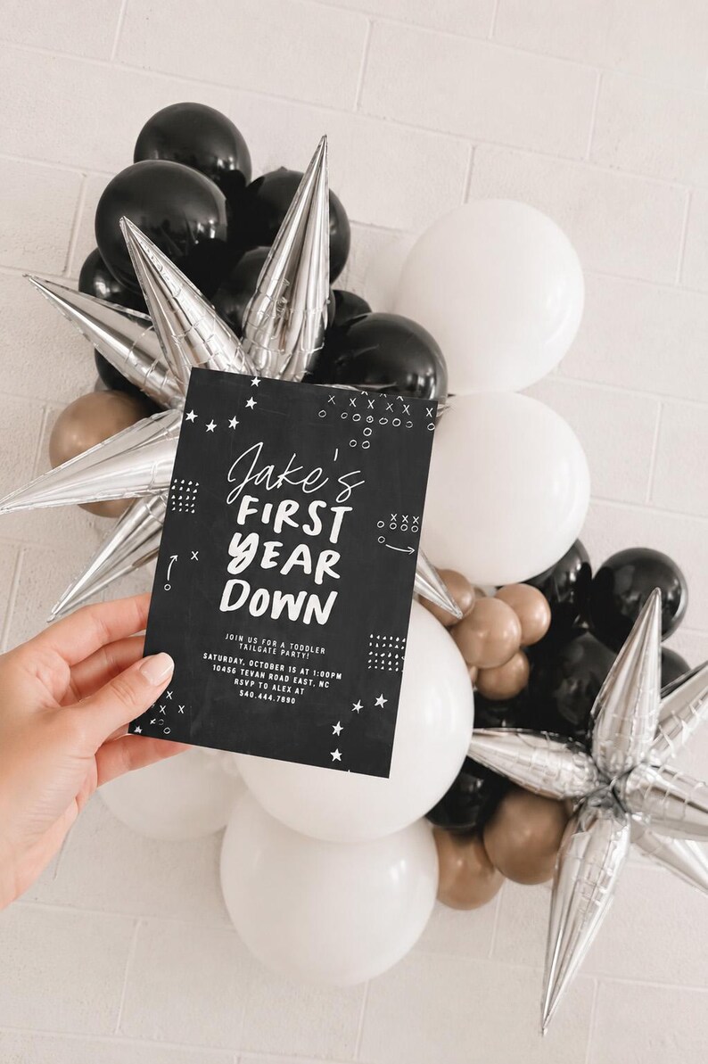 May include: A rectangular black invitation card with white text reading Jake's FIRST YEAR DOWN, including event details, set against a balloon arch featuring black, white, and metallic silver star-shaped balloons and small tan balloons.