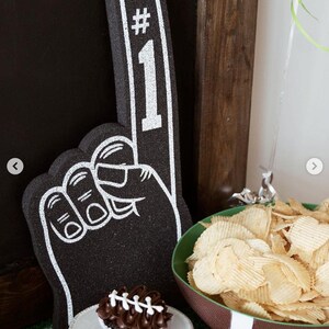 May include: A black foam finger with a white outline and the text #1 printed on the index finger. Next to it is a chocolate cupcake with white icing laces and a bowl shaped like a football filled with ridged potato chips on a green surface.