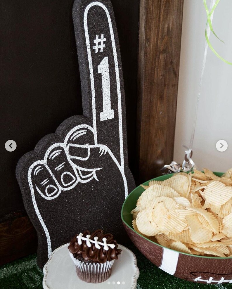 May include: A black foam finger with a white outline and the text #1 printed on the index finger. Next to it is a chocolate cupcake with white icing laces and a bowl shaped like a football filled with ridged potato chips on a green surface.
