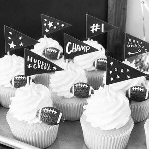 May include: A collection of cupcakes topped with white frosting, each decorated with a small textured football and a black triangular paper flag. The flags feature white printed text including #1, CHAMP, and HUDDLES + CUDDLES, alongside star and play diagram graphics.