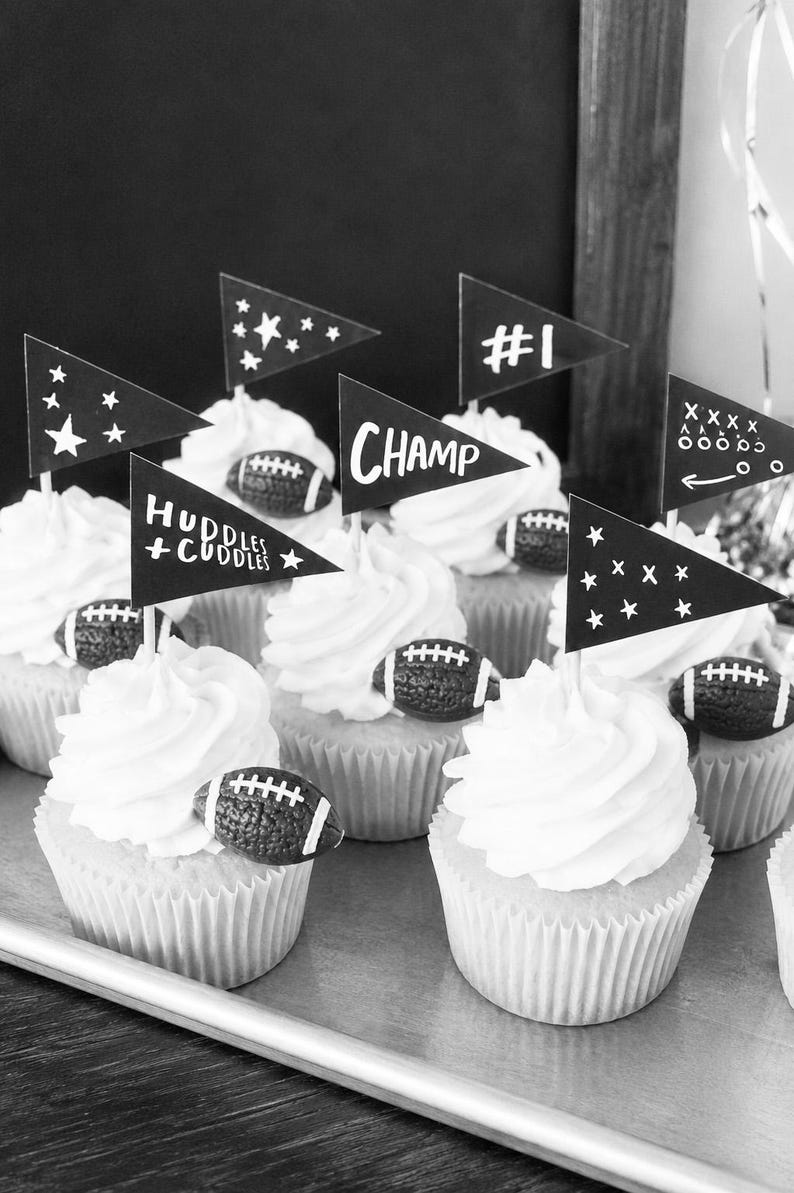 May include: A collection of cupcakes topped with white frosting, each decorated with a small textured football and a black triangular paper flag. The flags feature white printed text including #1, CHAMP, and HUDDLES + CUDDLES, alongside star and play diagram graphics.