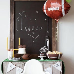 May include: A football-themed party display featuring a large chalkboard with white play diagrams, a brown football-shaped balloon, a white cake with a brown 'ONE' topper, a foam finger, a football-shaped bowl, and white pennant banners.