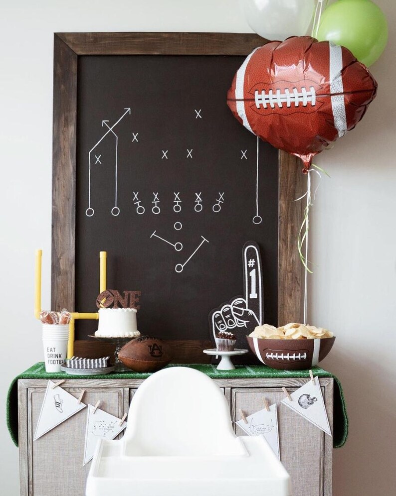 May include: A football-themed party display featuring a large chalkboard with white play diagrams, a brown football-shaped balloon, a white cake with a brown 'ONE' topper, a foam finger, a football-shaped bowl, and white pennant banners.