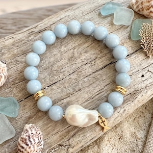 May include: A stretch bracelet featuring round, light blue beads, two gold-toned metal spacers, a single irregular white pearl, and a small gold-toned metal charm shaped like a fish with a cross symbol, arranged in a circular pattern.