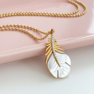 May include: A gold-toned chain necklace featuring a pendant shaped like a feather. The feather pendant is constructed with a gold-toned metal frame on one side and a white iridescent shell material on the other, accented by a central row of small stones.