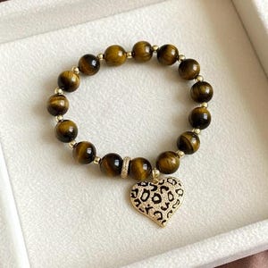 May include: A beaded bracelet featuring round, brown tiger's eye gemstones interspersed with small gold-toned metallic beads. The bracelet includes a central heart-shaped charm with a gold-toned border and a black and white leopard print pattern.