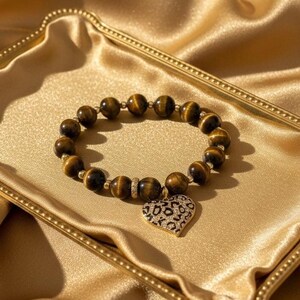 May include: A beaded bracelet featuring round, brown and gold tiger eye gemstones. The bracelet includes small gold-toned metal spacers and a single heart-shaped charm with a black and gold leopard print pattern suspended from the strand.