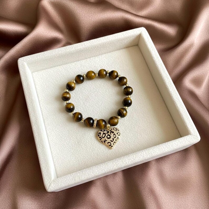 May include: A beaded bracelet featuring round, brown tiger's eye gemstones interspersed with small gold-toned metallic beads. The bracelet includes a central heart-shaped charm with a gold-toned border and a black and white leopard print pattern.