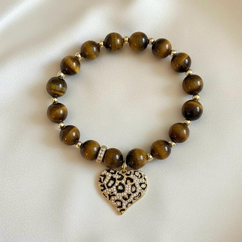 May include: A beaded bracelet featuring round brown tiger's eye gemstones interspersed with small gold-toned metal spacers. A single cylindrical spacer with embedded crystals sits near a heart-shaped pendant with a black and gold leopard print.