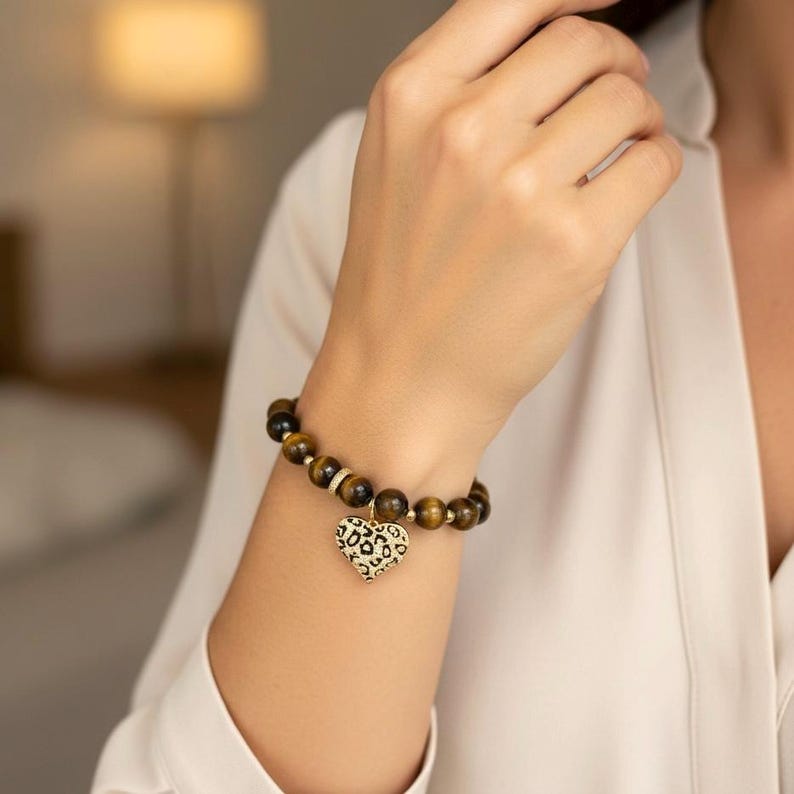 May include: A beaded bracelet featuring round brown tiger eye stones and small gold-toned spacers. A heart-shaped charm with a black and gold leopard print pattern hangs from the center of the bracelet, which is worn on a wrist.