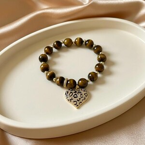 May include: A beaded bracelet featuring round brown tiger eye stones and small gold-toned metal spacers. A heart-shaped pendant with a black and white leopard print pattern and a pav&eacute;-set crystal border hangs from the center of the bracelet.