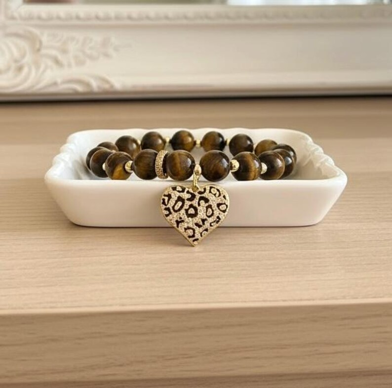 May include: A beaded bracelet featuring round brown tiger eye stones, small gold-toned spacer beads, and a central heart-shaped charm with a black and gold leopard print pattern. The bracelet is arranged in a circular shape on a white dish.