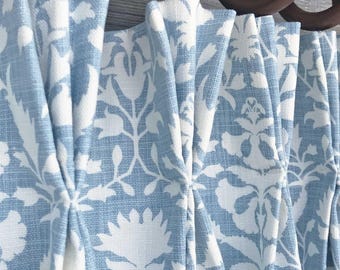 May include: A pleated curtain featuring a light blue and white floral pattern hanging from dark brown wooden rings on a rod. The fabric is labeled as Peacefulness in Luna. The curtain has deep vertical folds and a textured weave appearance.