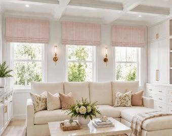 May include: Three light pink fabric Roman shades installed over three windows. The shades feature a soft, textured weave and are positioned above a cream-colored sectional sofa with various decorative throw pillows and a wooden coffee table.