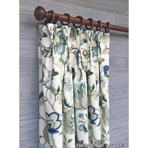 May include: A pleated curtain panel featuring a cream background with a floral pattern in shades of blue, green, and tan. The curtain hangs from a dark brown wooden rod with matching rings. The fabric is labeled as Brissac in Sapphire.
