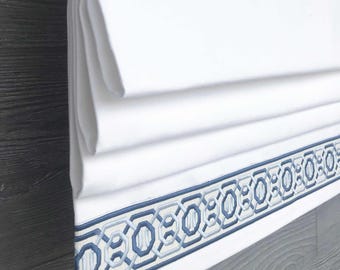 May include: A white fabric roman shade featuring horizontal folds and a decorative border at the bottom. The border displays a repeating geometric pattern embroidered in shades of dark blue and light blue, creating a structured, linear design.