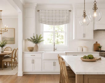 May include: A kitchen interior featuring white shaker cabinets, a farmhouse sink, and a window with a beige and white plaid roman shade. A large kitchen island with a white countertop sits in the foreground, complemented by two glass pendant lights.