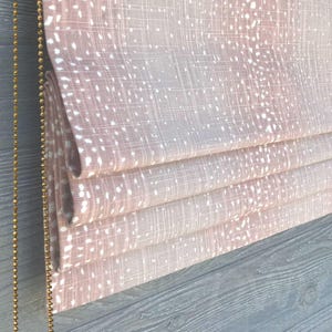 May include: Close-up of a folded, light pink roman shade with a white speckled pattern. A gold beaded chain hangs on the left side. The shade is made of a textured fabric and is partially folded.