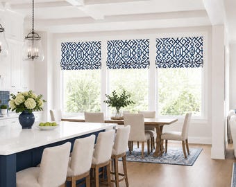 May include: Three roman shades featuring a navy blue and white geometric pattern installed over three large windows. The shades are constructed with fabric folds and are positioned above a dining area with light-colored chairs and a wooden table.