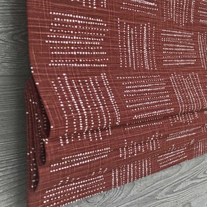 May include: Close-up of a folded, rust-colored fabric with a repeating pattern of white dotted lines. The lines are arranged in a grid-like design, creating a textured appearance. The fabric is likely used for home decor, such as curtains or blinds.