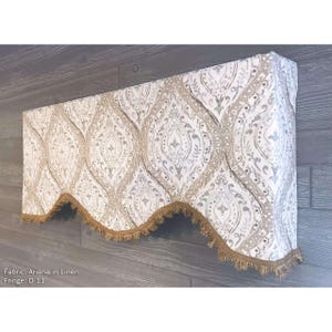 May include: A window valance featuring the Ariana in Linen fabric pattern, characterized by a repeating beige and white damask-style motif. The bottom edge is finished with a decorative brown fringe trim, creating a scalloped silhouette.
