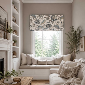 May include: A window shade featuring a floral pattern in shades of blue, brown, and cream. The fabric is folded in a horizontal Roman style, covering the upper portion of a window that overlooks a green outdoor area with trees.